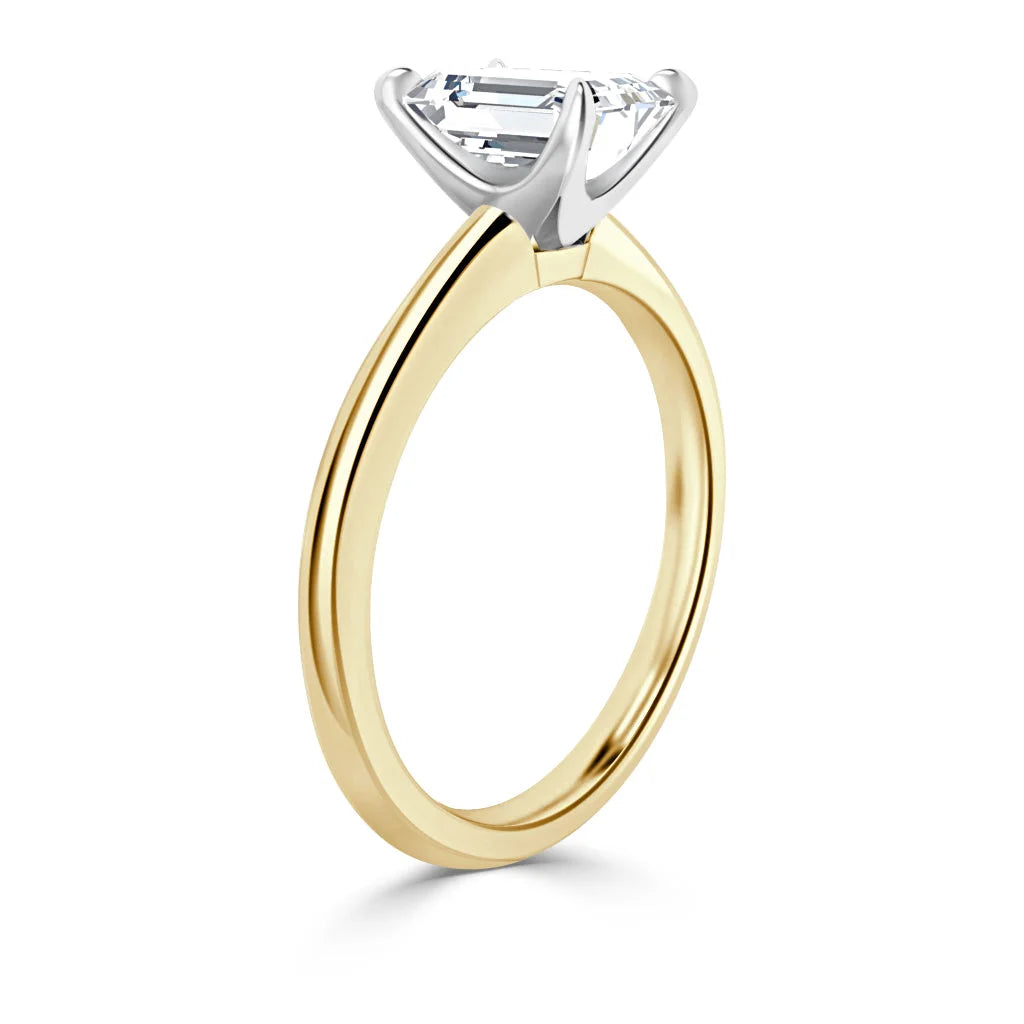 A gold ring with a solitaire emerald-cut diamond set in a white gold prong setting, viewed from the side.