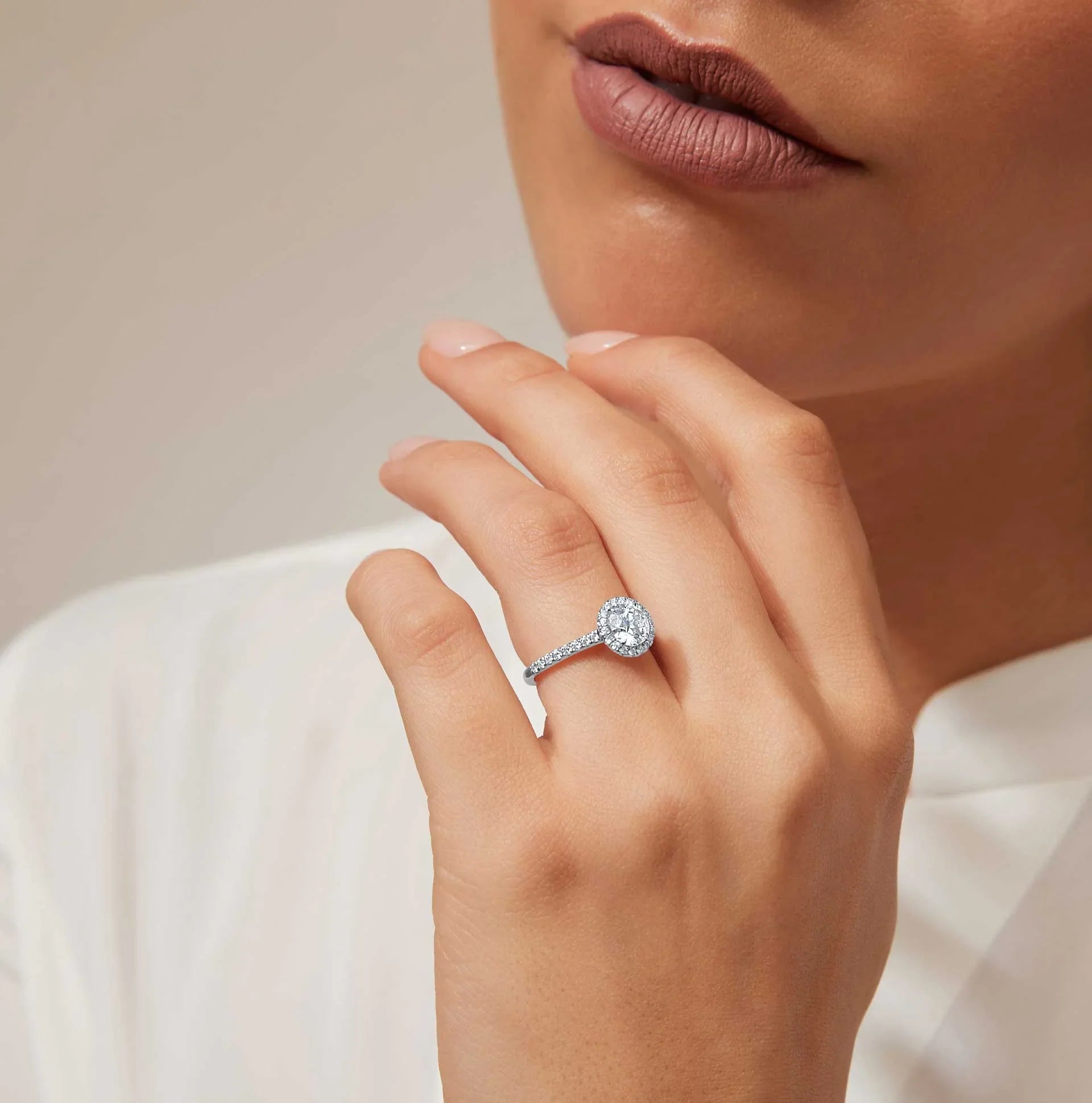 A woman wearing a diamond halo engagement ring on her left hand, with neutral lipstick and a white blouse.