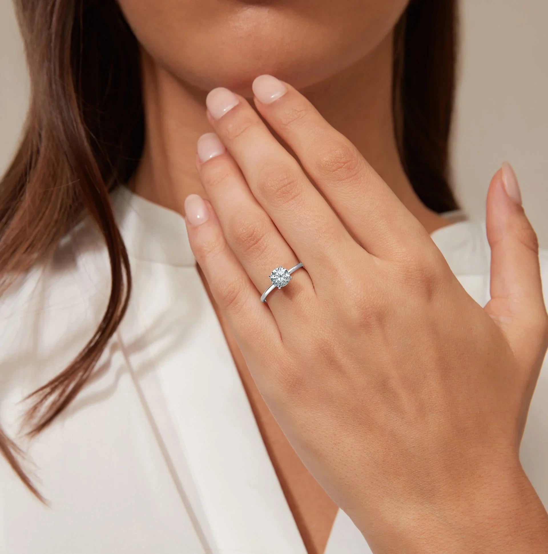A woman with light pink nails wears a silver solitaire diamond ring on her left hand, touching her face.