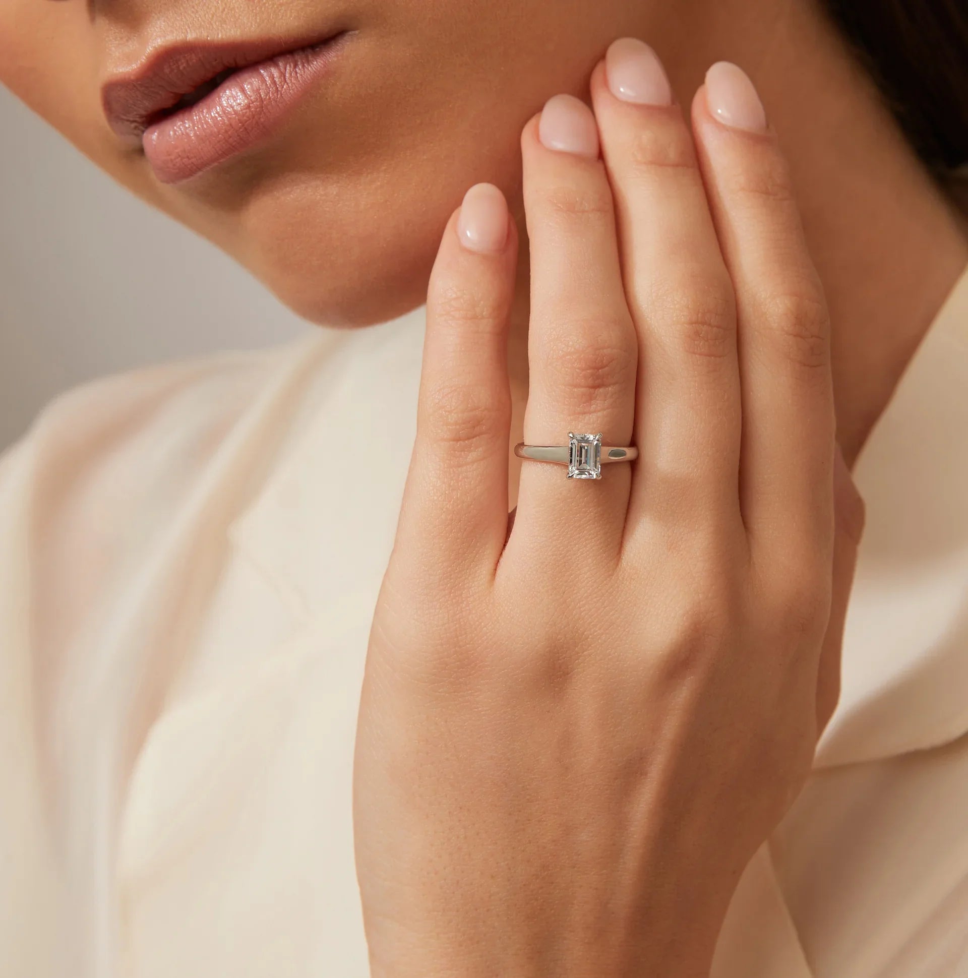 Woman with manicured nails wearing an emerald-cut diamond ring on her finger, hand near her face.
