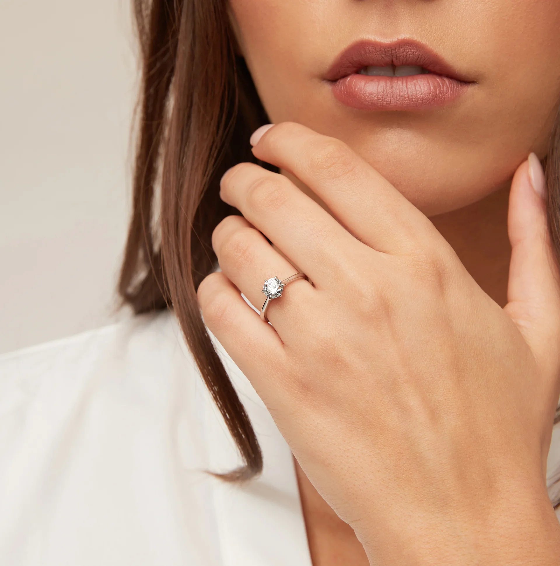 Woman with neutral lipstick wears a solitaire diamond ring on her left hand, touching her face.