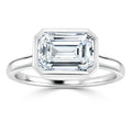 A silver ring features a large emerald-cut diamond with a sleek, geometric bezel setting on a plain band.