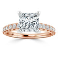 Rose gold ring with a large princess-cut diamond and small round diamonds on the band, on white background.